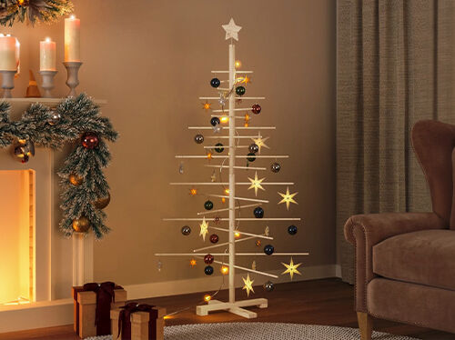 ミニマルな木製クリスマスツリー。オーナメントと星で飾られ、暖炉やラッピングされたギフト、ブラウンのアームチェアの横に置かれているリビングルームの床に配置。コレクションの商品を見るにはクリックしてください。