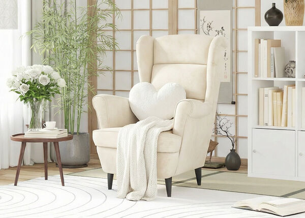 クリーム色のアームチェアにハート型のクッションとスローブランケットを添え、小さな木製のサイドテーブルには白いバラとカップを配置。竹の観葉植物、間仕切り、そして本でいっぱいの白いキューブ型収納ユニットを背景にした、スタイリングされたリビングルーム。