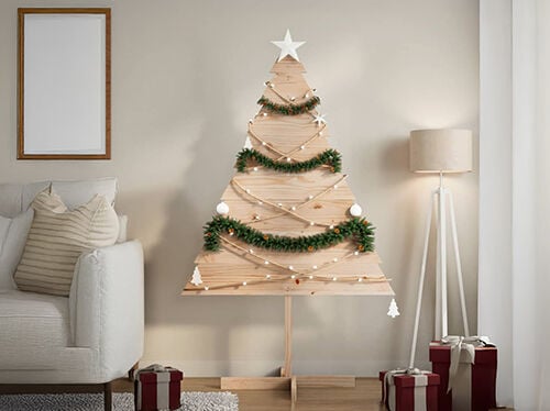 パネル製の背の高い木製クリスマスツリーが、ガーランドとイルミネーションで飾られ、ソファとフロアランプのそばに置かれています。床にはラッピング済みのギフトが並んでいます。コレクションの製品を見るにはクリックしてください。