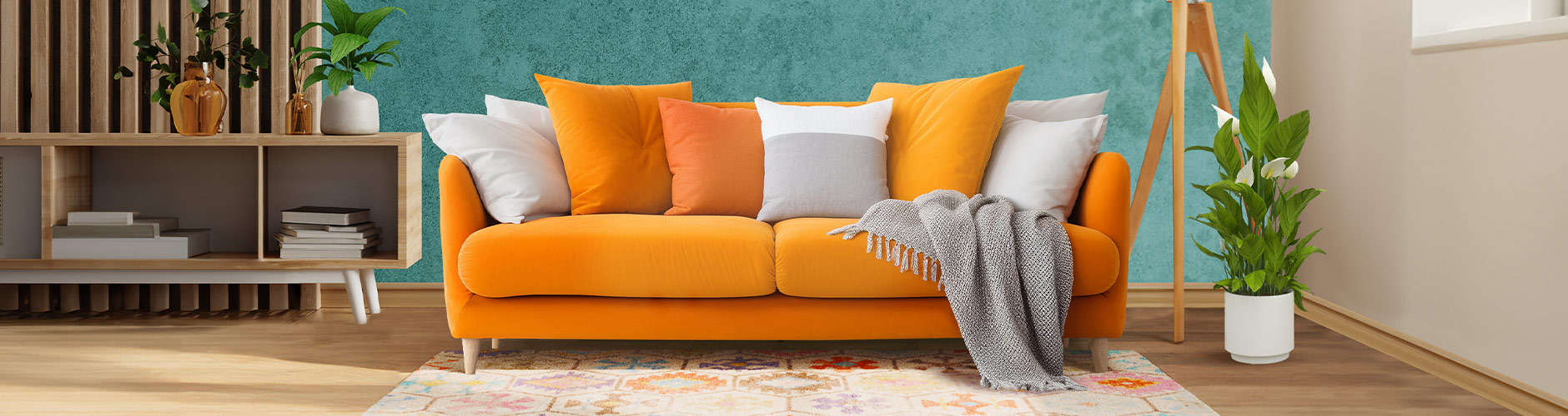 オレンジ色のソファにオレンジと白のクッションとグレーのブランケットが置かれたリビングルーム。ソファの下にはカーペットが敷かれ、横には緑の観葉植物。オープンシェルフのテレビ台には本が並び、上にはいくつかの花瓶が置かれている。